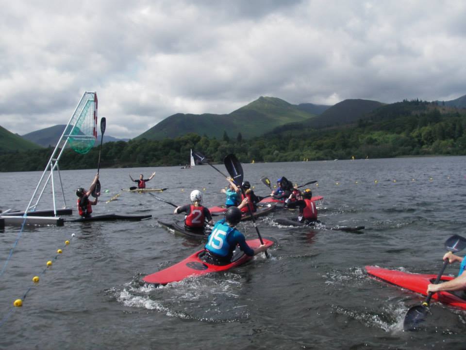 Canoe polo players approaching a goal on a lake with wooded hills behind them.