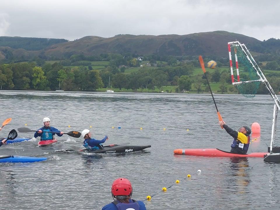 A canoe polo goalkeeper reaching up with a paddle to stop a shot on the lake.
