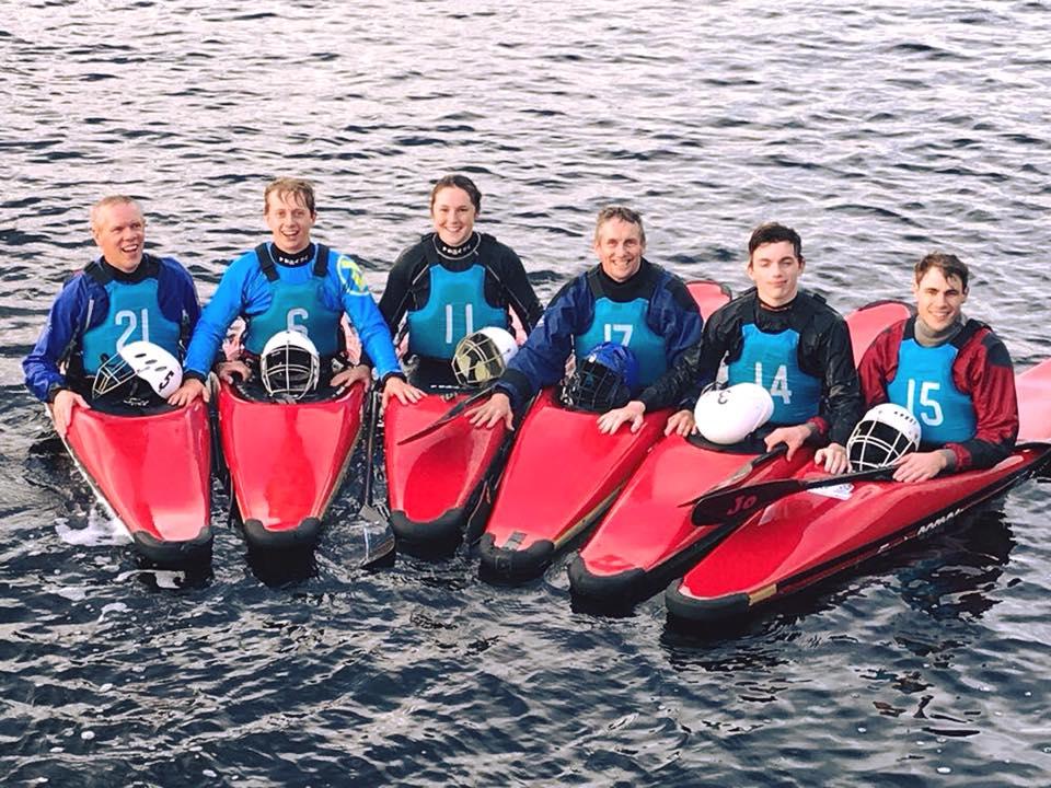 Six paddlers smiling from a row of red kayaks on open water.