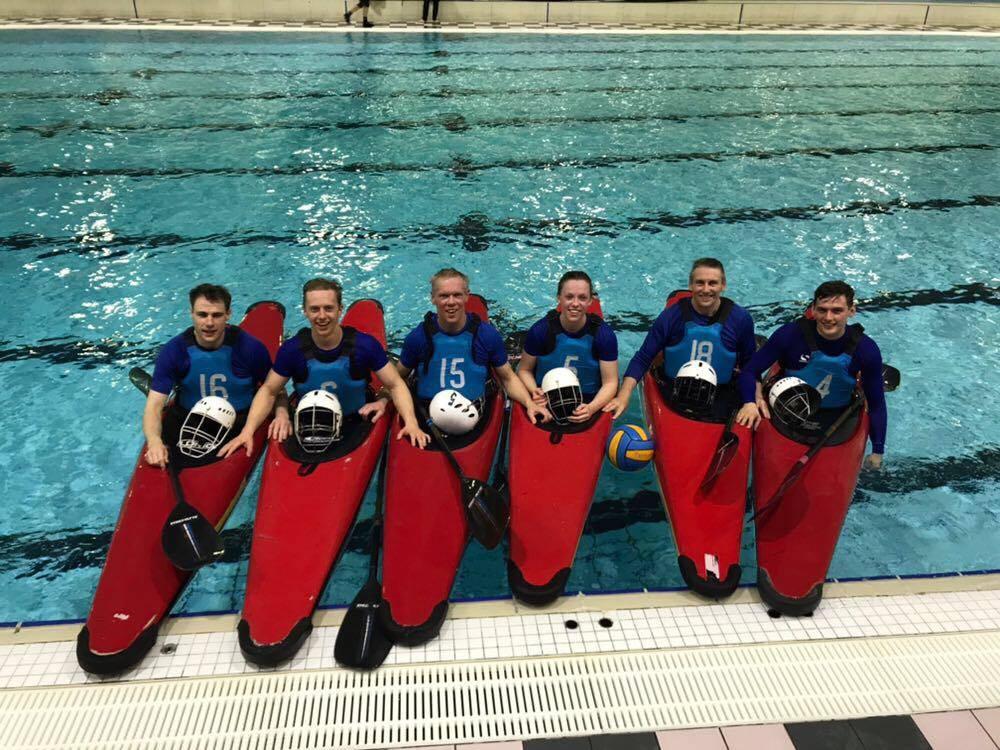 Six paddlers in blue buoyancy aids lined up in red kayaks at the poolside.