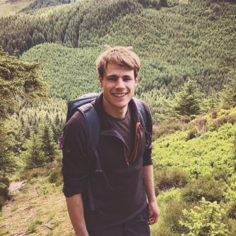 A smiling walker on a hillside path above a forest.