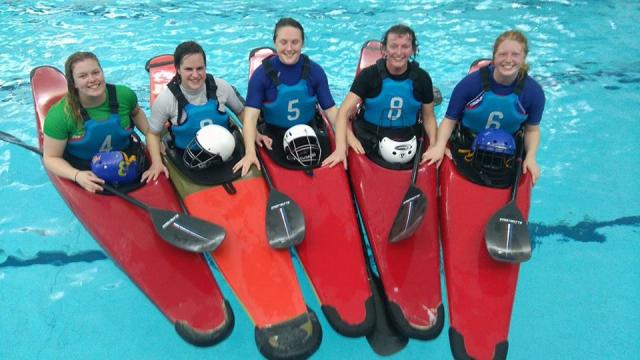 Five paddlers lined up in red kayaks for a poolside team photograph.