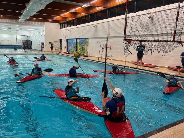 Players contesting the ball below a canoe polo goal in an indoor pool.