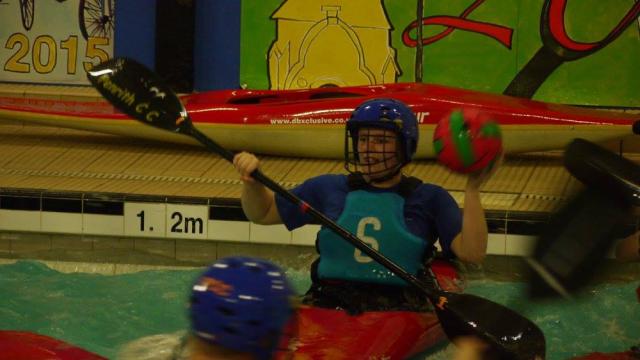 A canoe polo player holding a ball and paddle in an indoor pool.